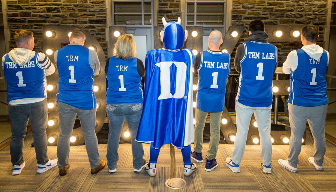 TRM Labs team members and the Duke Blue Devil mascot showing the backs of their branded Duke jerseys